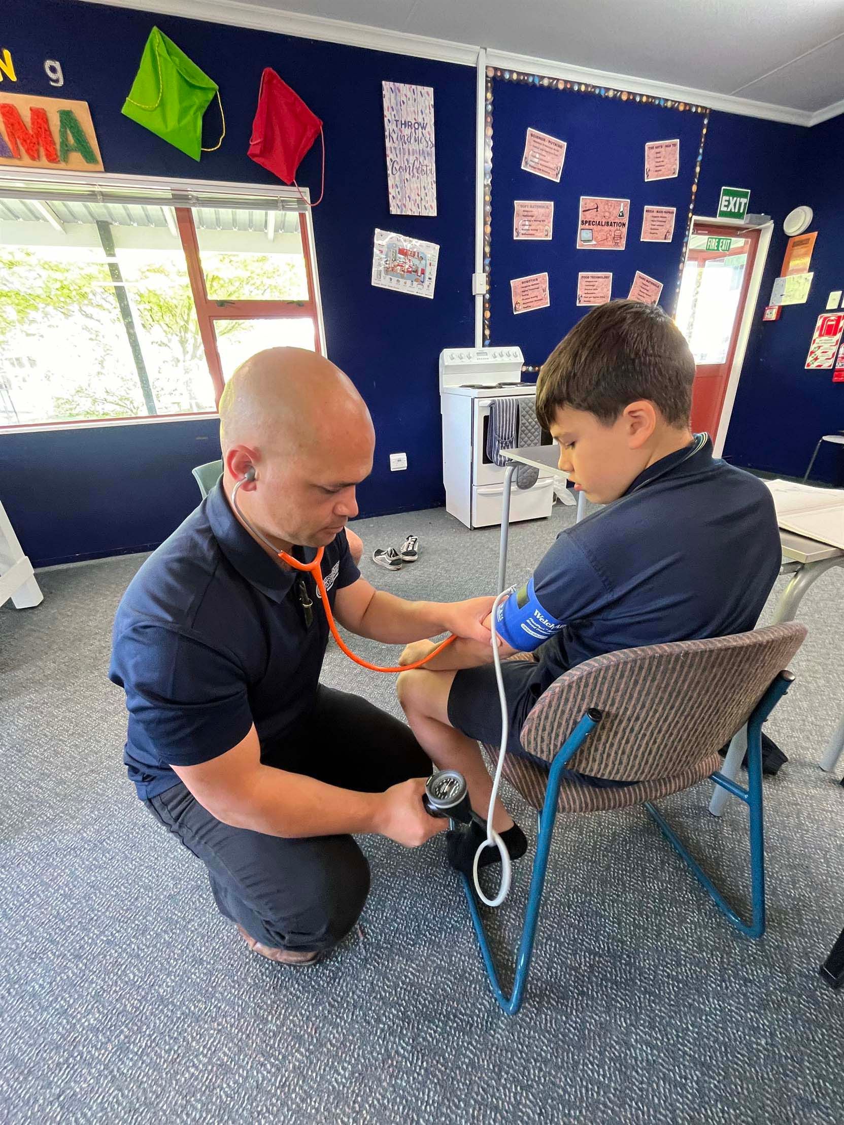Josh Te Rauna kneeling beside a boy in a school classroom, taking a blood pressure reading with a stethoscope and cuff.