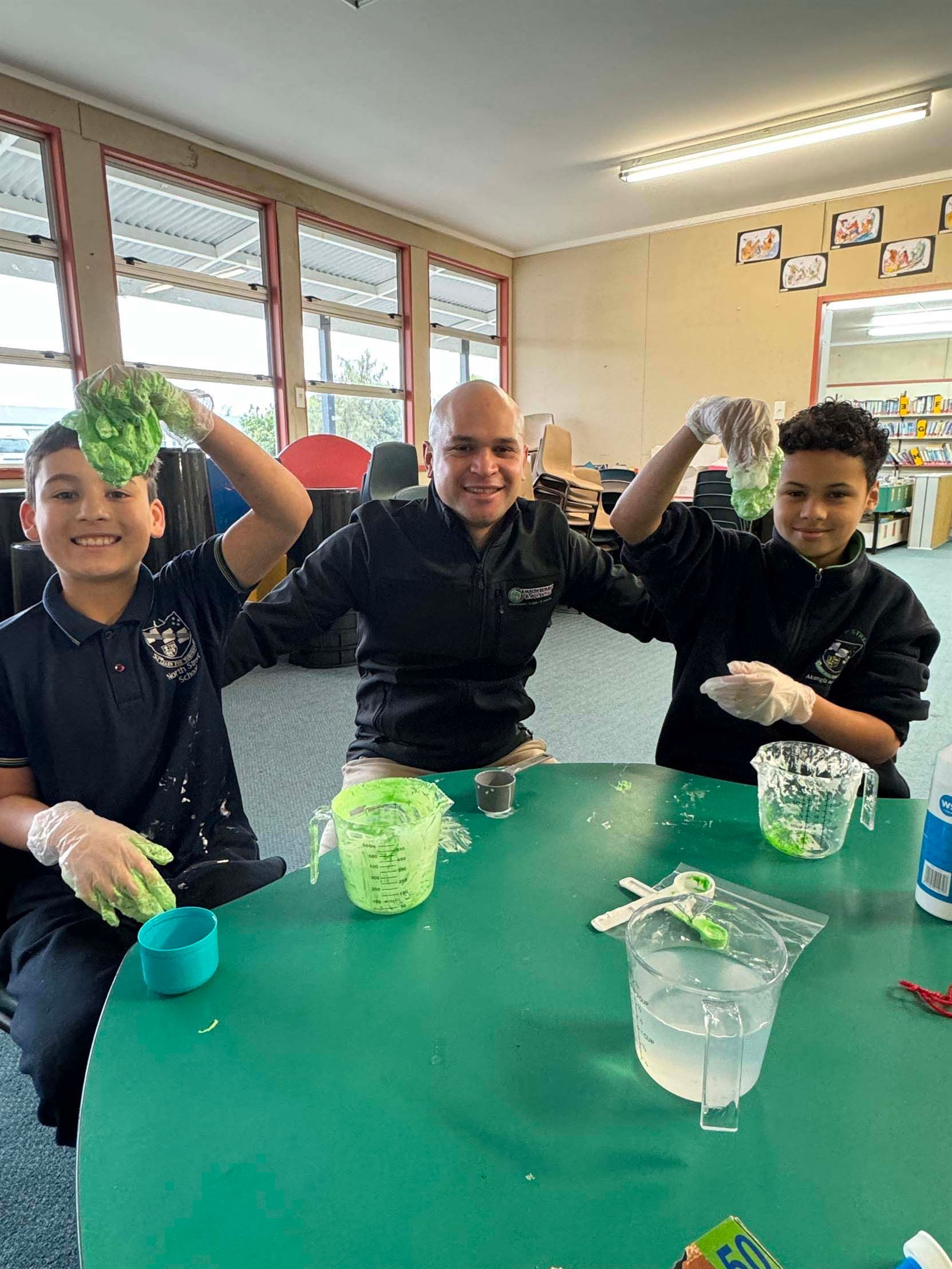 Josh Te Rauna working with two boys on a hands-on activity in a school classroom.